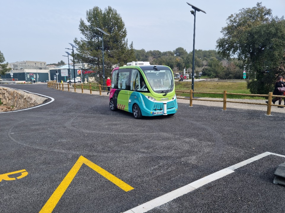 A photo of a green and blue Navya autonomous shuttle on a test track with a person in the background, surrounded by trees and a parking area.